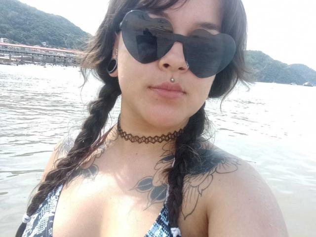 DaisyFx with black braids, nose ring, black choker, tattoos on shoulders, large black sunglasses, and blue-checkered bikini top, standing in water with mountains in the background.