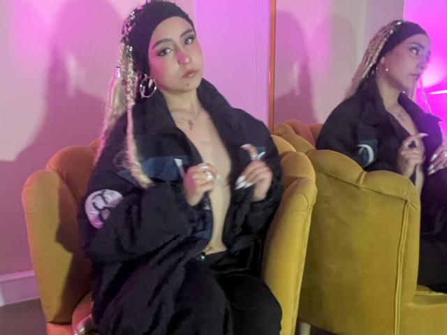 MaveParker with braided hair, hoop earrings, and black jacket, partially open to reveal small tits, sitting on yellow chair, holding a drink, reflected in mirror.