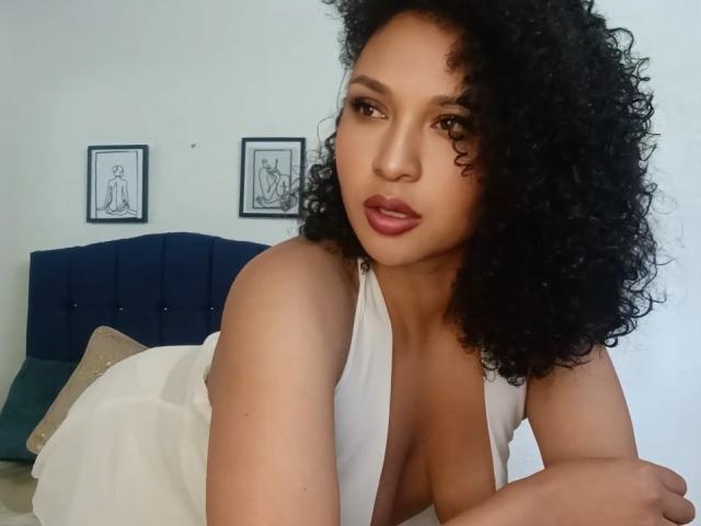 CarlaMuille with curly black hair, brown eyes, and full lips, wearing a white dress with a deep neckline, showing her boobs, leaning on a bed with blue headboard.