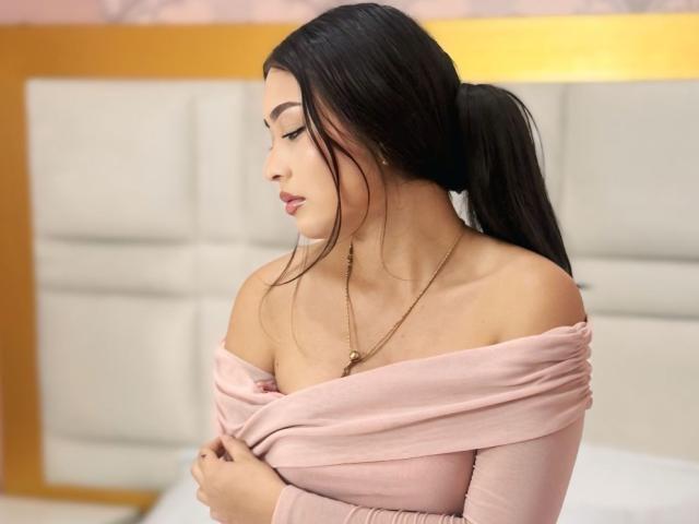 BellaMiaI with long black hair in a ponytail, wearing a pink off-shoulder top, small gold necklace, looking down, arms crossed, showing her tits.