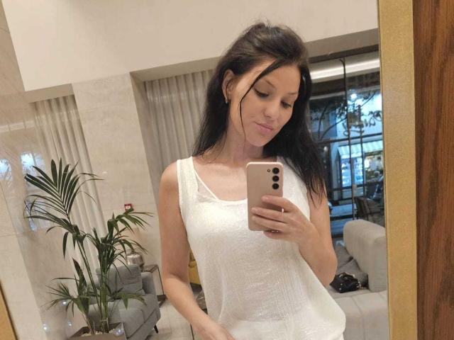 JoliDanielle with long black hair, white sleeveless top, and subtle smile, holding a phone, taking a mirror selfie, showing small tits, in a modern, well-lit room.