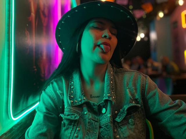 EmiliaCartersh with long black hair, green neon light, tongue out, wearing a studded denim jacket and black cowboy hat, showing a tattoo on her neck.
