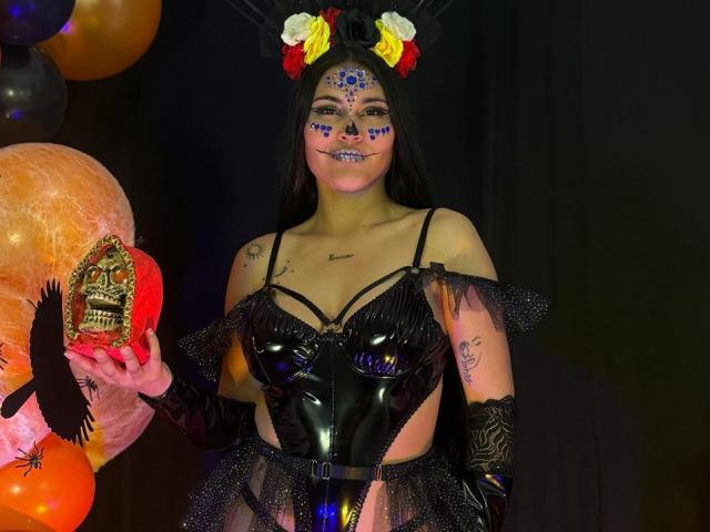 EmiliaCartersh in shiny black latex lingerie, holding a skull, face painted with colorful sugar skull design, wearing flower headpiece, long black hair, black gloves, and tattoos.