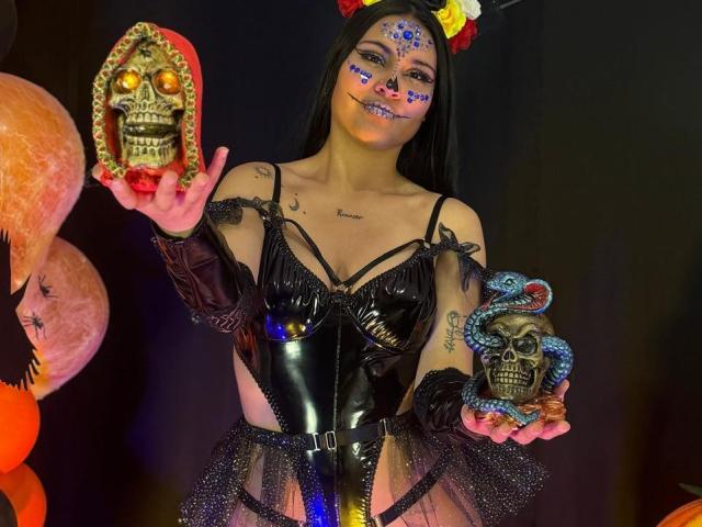 EmiliaCartersh with black latex outfit, cat ears, face paint, holding skull masks, tattoos, long black hair, black gloves, black lace, and blue face paint on dark background.