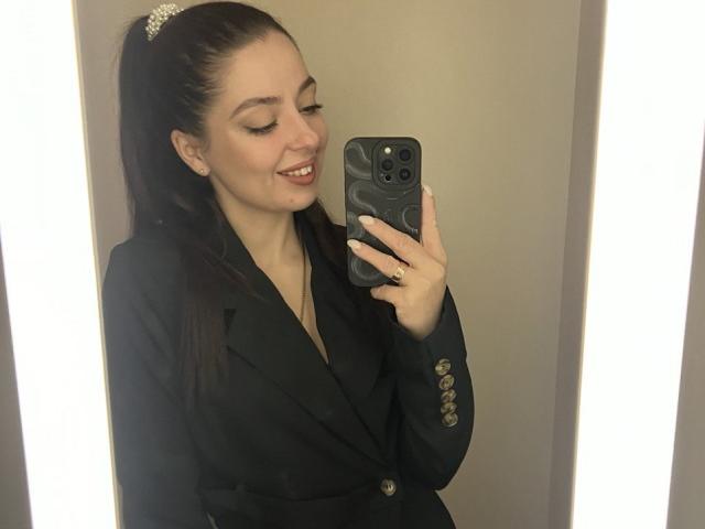 DiamondEyesss, with long brown hair in a ponytail, wearing a black blazer with gold buttons, smiling at a mirror selfie holding a black phone. Boobs not visible.