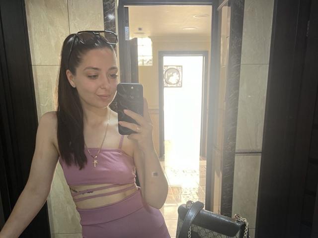 DiamondEyesss with long brown hair in a ponytail, wearing a pink sports bra and matching shorts, taking a mirror selfie, showing small tits and a subtle tattoo on her arm.