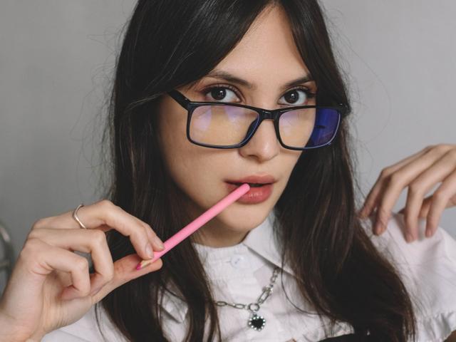 MiraPerez with long dark hair, blue-framed glasses, white shirt, holding pink lipstick to her slightly parted lips. She has a necklace with a black flower.