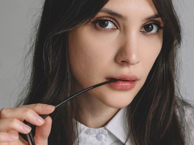 MiraPerez with dark brown hair, holding a black pen to her slightly parted lips, looking directly at the camera with big brown eyes. Her skin is fair and smooth.