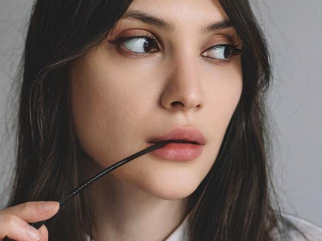MiraPerez with straight brown hair, light skin, and brown eyes, holding a black eyeliner pencil to her slightly parted lips. Her eyeliner is subtle, enhancing her natural look.