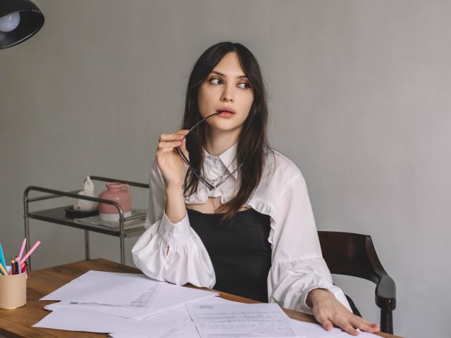 MiraPerez with long dark hair, white shirt, black dress, holding pen to her lips, papers on wooden desk, black lamp, metal shelf with pink box, and pencils in front.