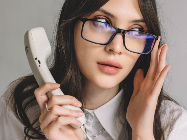 MiraPerez with glasses, holding a white landline phone, her hand near her ear, wearing a white collared shirt, looking slightly to the side with a subtle smile.