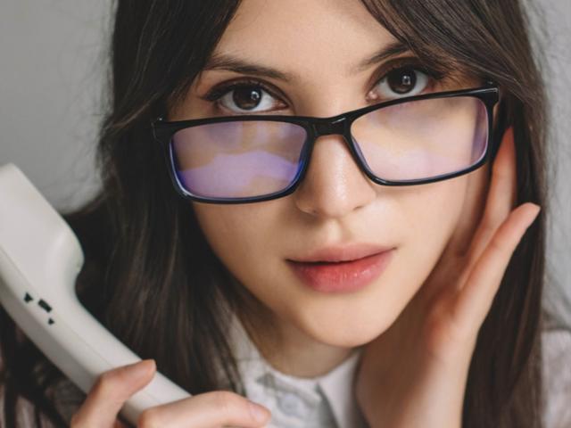 MiraPerez with glasses, holding a white phone to her ear, brown eyes, long brown hair, pink lips, white shirt, hand on cheek, looking slightly to the left.