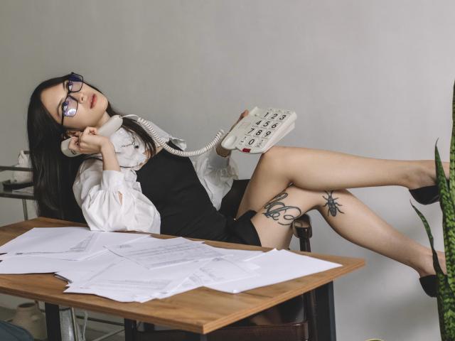 MiraPerez with glasses, black dress, white shirt, talking on a landline, legs up, tattoo on thigh, papers on desk, black heels, white wall, green plant.
