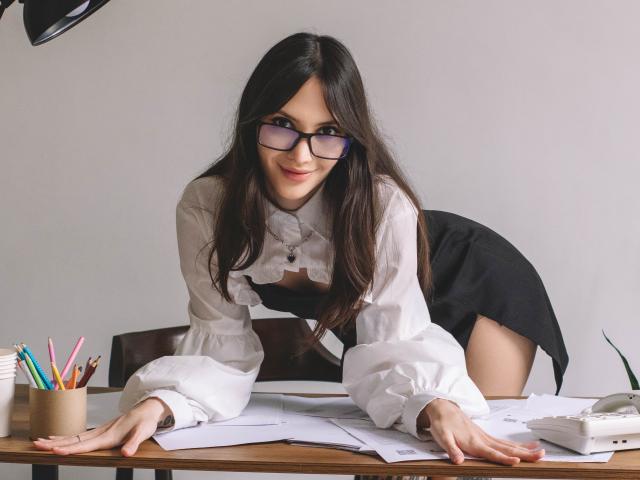 MiraPerez with long brown hair, glasses, white ruffled shirt, black skirt, leaning on desk, smiling, hands on papers, pencils in cup, phone on right, wooden chair behind.