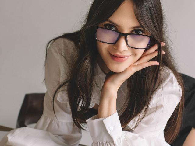 MiraPerez with long brown hair, wearing glasses and a white shirt, leaning on her hand with a subtle smile, showing her cute face and small, pretty tits.