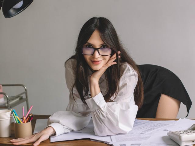 MiraPerez with long dark hair, glasses, white shirt, black skirt, leaning on desk, smiling, hand on cheek, pens in cup, papers, and desk lamp in a modern office.