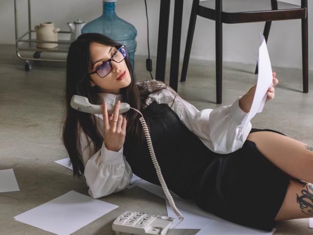 MiraPerez with glasses, lying on floor, black dress, white shirt, holding phone, papers around, tattoo on thigh, blue water bottle in background, wooden chair to the right.