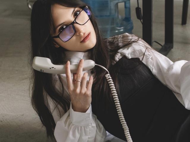 MiraPerez with glasses, holding a white corded phone to her ear, wearing a black dress and white shirt, leaning forward slightly. She has long black hair and a ring.