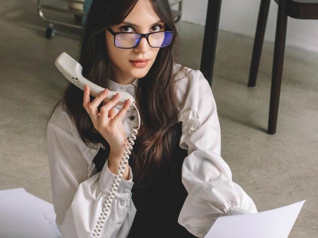 MiraPerez with glasses, holding a white landline phone, wearing a white shirt and black dress, papers in front, long dark hair, sitting, slightly smiling, concrete floor.