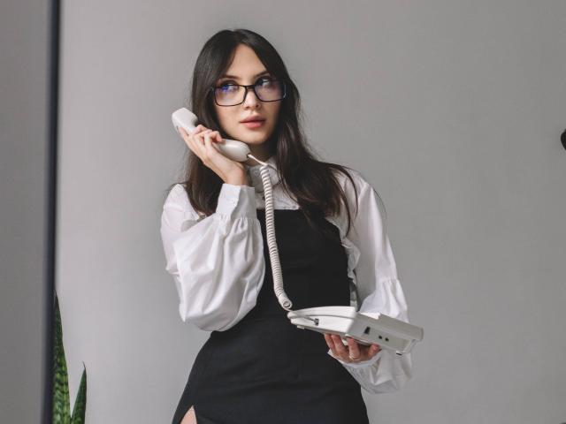 MiraPerez with glasses, long dark hair, white shirt, black dress, holding phone to ear, holding white binder, standing, grey background.