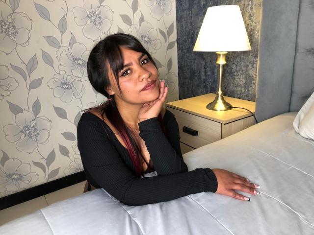 ArianaCruz, with dark hair in pigtails, leans on a bed, wearing a black ribbed top, exposing her small tits, hand on chin, lamp on wooden nightstand, looking at camera.