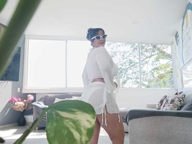 TamaraFoxy with white mesh top and shorts, white sunglasses, standing, showing her ass, dark hair in bun, in bright room, green plant in foreground, sunlight.