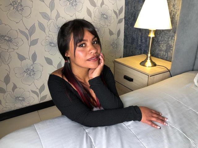ArianaCruz with dark hair, small tits, wearing a black ribbed top, hand on chin, sitting on a bed, lamp on wooden nightstand, white quilt, floral wallpaper.