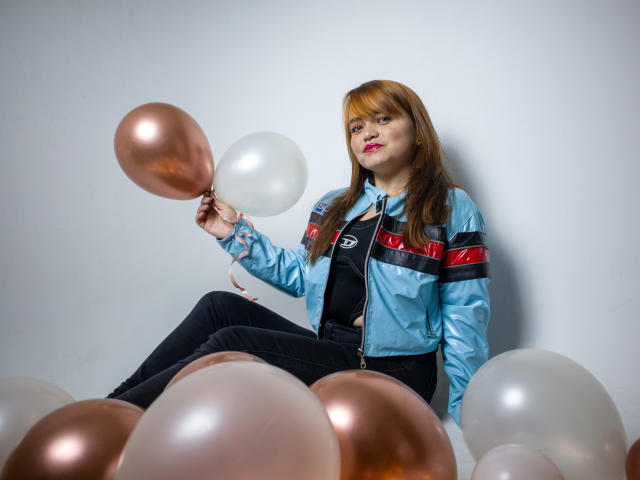 MoonRay69, avec du rouge à lèvres, assise parmi des ballons cuivrés et blancs, tenant un ballon cuivré, portant une veste bleue brillante, un t-shirt noir et un pantalon noir, avec un regard mignon et confiant..