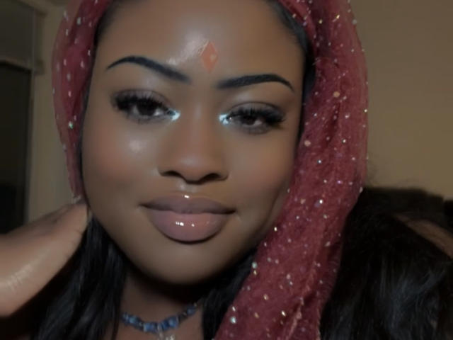 Le visage de MinaBlu, avec un foulard rouge scintillant, une peau brun foncé, des lèvres brillantes et un petit point rouge sur le front, regardant légèrement sur le côté avec un sourire doux..