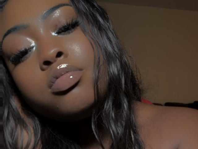 Il viso marrone lucido di MinaBlu, con le labbra rosa brillanti, i lunghi capelli neri ondulati e le ciglia drammatiche, con gli occhi chiusi e la testa leggermente inclinata all'indietro, appare sexy e attraente..
