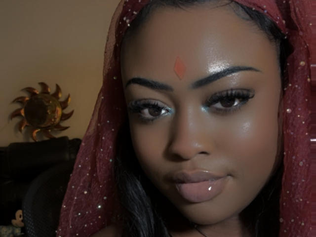 MinaBlu has dark brown skin, black hair, and wears a red headscarf with sparkles. She has a red bindi on her forehead, glossy lips, and long eyelashes.