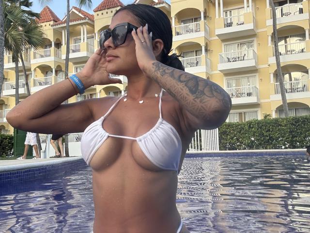 ThaliaAres, con tatuaggi sul braccio, indossa un bikini bianco e sta sistemando gli occhiali da sole vicino alla piscina. Ha un seno abbondante ed è in piedi in acqua blu..