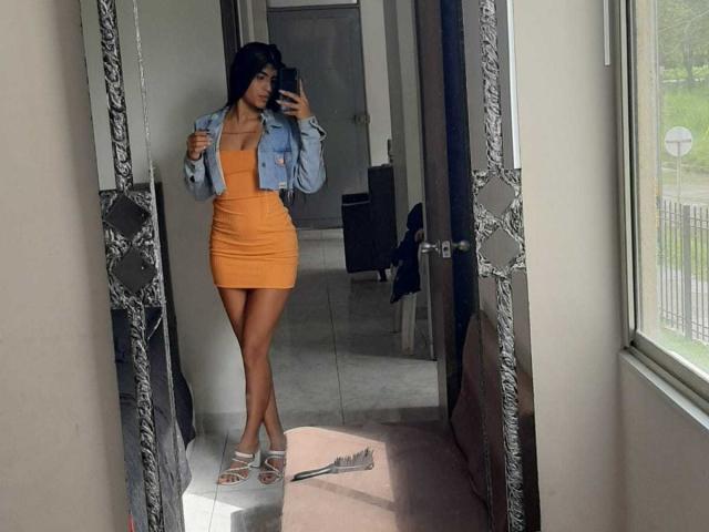 JulieTeal with long black hair, wearing an orange dress, denim jacket, and white sandals, standing in front of a mirror, holding a phone, showing her ass and tits.