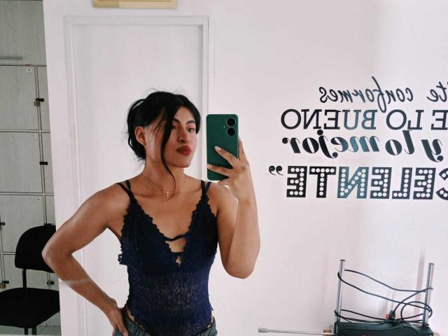 MichelleDenise with dark hair in a messy bun, wearing a black lace bodysuit with cutout, holding a green phone, standing with hands on hips, showing her tits.