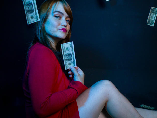 MoonRay69 portant un pull rouge, tenant des billets de dollar, assise, les jambes croisées, lumière bleue, rouge à lèvres rose, sourire narquois, cheveux bruns, légère rougeur sur les joues, argent en arrière-plan, pose détendue..