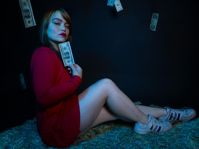 MoonRay69, vêtue d'une robe rouge, tenant de l'argent liquide, assise sur de l'argent, avec des billets de dollar flottant autour, portant des baskets blanches et rouges, éclairage bleu et rouge, avec une expression satisfaite..
