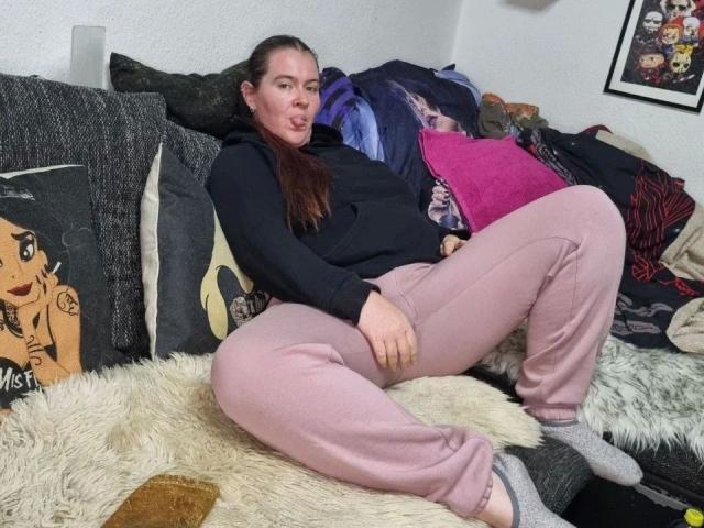 Rotkaeppchen-hot with brown hair, puffy pink pants, and black hoodie, sitting on a furry blanket, sticking her tongue out, legs spread, gray socks, messy pillow background.