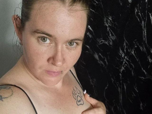 Rotkaeppchen-hot, topless with tattoos on shoulders, has a nose ring and a lip piercing, brown hair in a messy bun, holding straps, black and white background.
