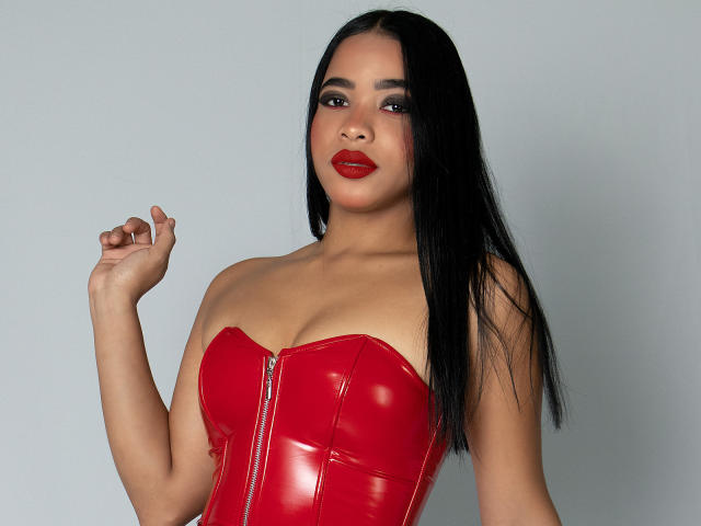 KarlaBella with long black hair, red glossy leather corset, and red lipstick, standing, right hand raised, looking at camera, showing her tits and sexy face.