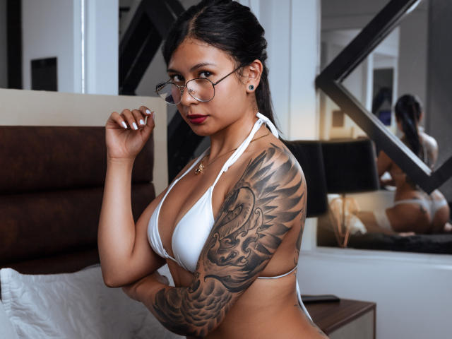 JessicaConti with glasses, white bikini, and tattoos on her right arm, showing off her tits and ass, standing by a bed, looking over her shoulder with red lipstick.