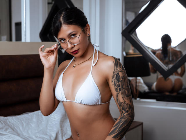 JessicaConti with glasses, white bikini top, tattooed arm, brown skin, red lipstick, holding glasses, mirror reflection shows her butt, bedroom, bed with white sheets, brown headboard.