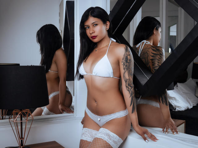 JessicaConti with tan skin, black hair, white bikini, white panties, lace thigh-highs, tattoos on right arm, sitting on bed, reflection in mirror, looking at camera, red lipstick.