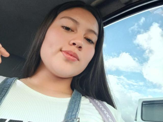 NatalliieSweet, con capelli neri lisci, pelle chiara e labbra rosa, indossa una tuta di jeans e una maglietta bianca a coste, e sorride leggermente seduta in un'auto..