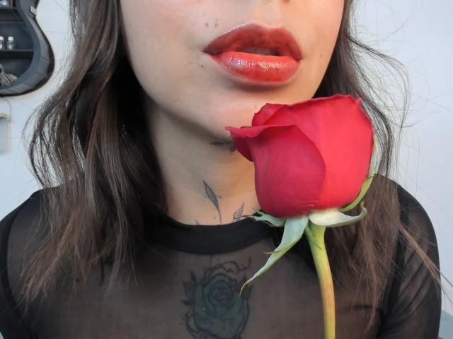 KayleeM's shiny red lips are slightly parted, holding a red rose to her lower lip. She's wearing a sheer black top, revealing a rose tattoo on her chest.