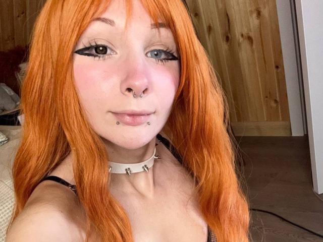 HannaCool with orange hair, black eyeliner, nose piercings, lip piercings, and a spiked choker, wearing a black top, in a selfie. Her tits are small.