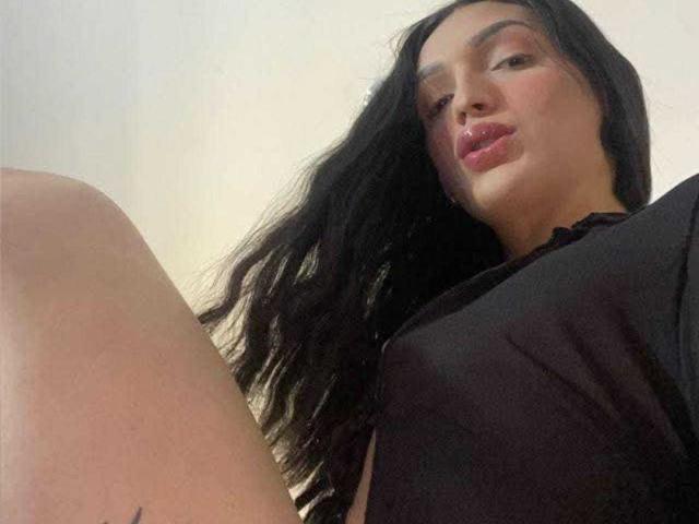 isabellaSweettx with black hair, pouty pink lips, wearing a black top, legs up, slight tan, looking down at camera, soft light, subtle shadow.