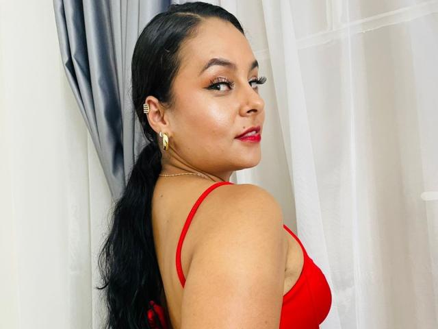 LunaSoler with dark hair in a ponytail, wearing a red bra, gold earrings, and red lipstick, looking over her shoulder with a slight smile, showing her ass.