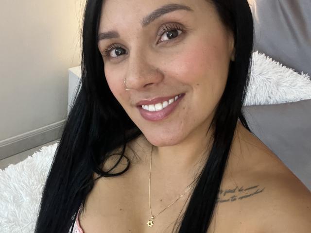 YessiJaymes tem cabelos pretos longos, olhos castanhos, um piercing no nariz, um sorriso que mostra dentes brancos e uma tatuagem no ombro direito. Ela está usando um colar com um pingente em forma de estrela..