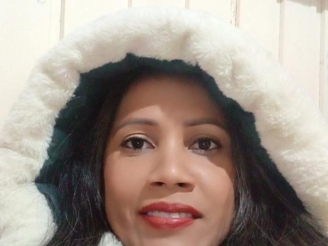 NoemiBelle with dark hair, brown eyes, and red lipstick, wearing a fluffy white hood, smiling softly. Her nose is centered, and her skin is smooth and tan.