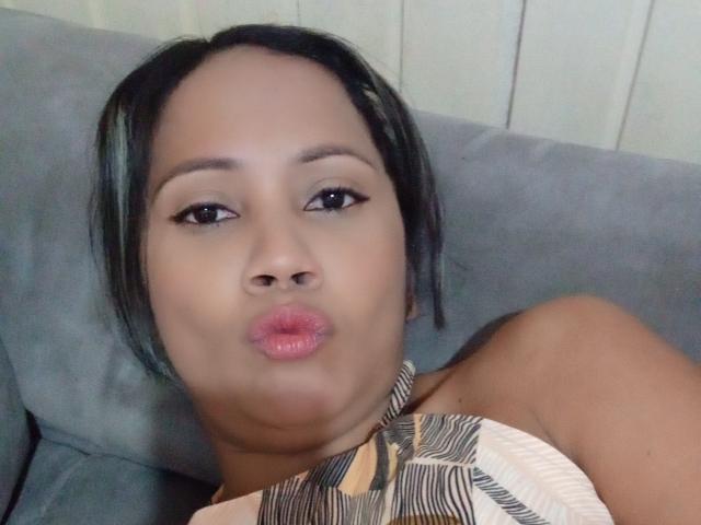 NoemiBelle has dark brown skin, black straight hair, and is puckering her pink glossy lips. She's wearing a patterned top and lying on a grey couch.